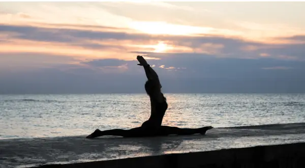 Silhouette di una donna in Ardha Hanumanasana sulla riva del mare al tramonto, con le braccia alzate verso il cielo.