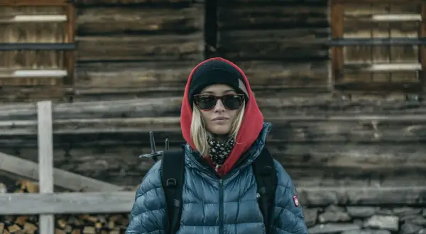 Kobieta w czerwonej bluzie z kapturem i czarnej czapce, w okularach przeciwsłonecznych, z plecakiem, przed drewnianą chatą.