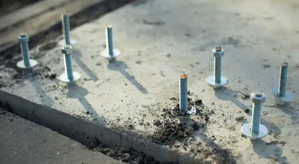 Kilka metalowych kotew z podkładkami i nakrętkami osadzonych w betonie.