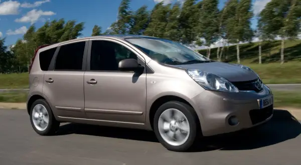 Złoty Nissan Note 2010 pędzi po drodze, drzewa na tle falują na wietrze.