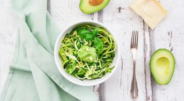 Zoodles con pesto di avocado, guarniti con pinoli e basilico. Accanto, mezzo avocado, un pezzo di parmigiano e una forchetta.
