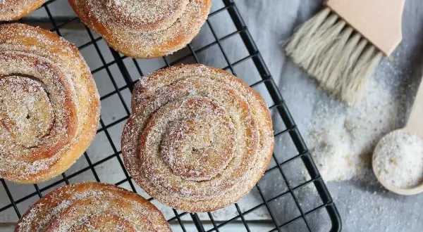 Nybakade kanelbullar med strössel av brunt socker och pärlsocker på ett svart galler. En träpensel och en sked med socker ligger bredvid.