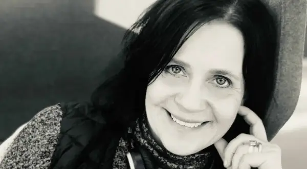 Karin Ståhl ler mot kameran, med handen vid kinden. Hon bär hörlurar runt halsen och en väst över en mönstrad tröja.