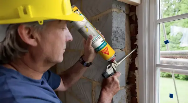 Hombre con casco amarillo usa sellador de poliuretano para rellenar huecos en instalación de ventana.