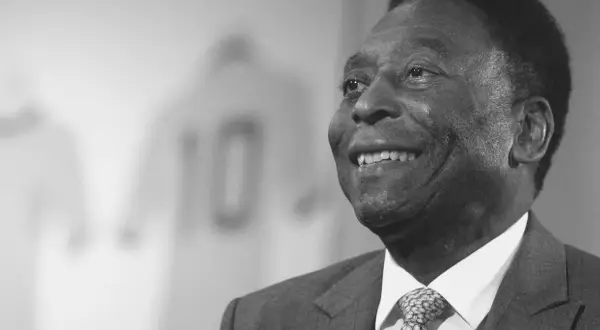Uśmiechnięty Pele, legenda futbolu, w garniturze. W tle koszulka z numerem 10. Czy Pele żyje? Tak, jego dziedzictwo żyje wiecznie.