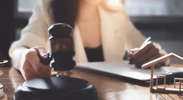 Sądowy młotek i model domu symbolizują licytację komorniczą. Kobieta podpisuje dokumenty, przygotowując się do sprzedaży nieruchomości.