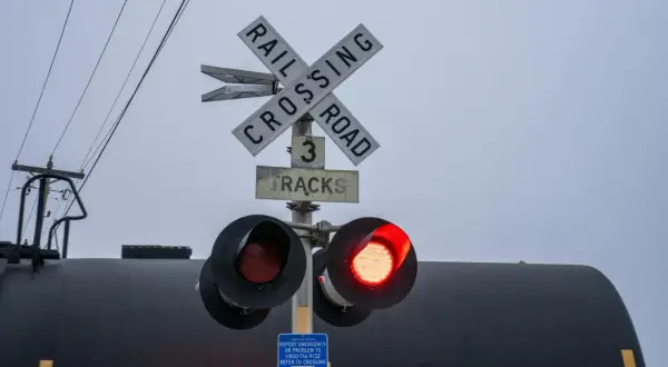 Przejazd kolejowo-drogowy z czerwonym światłem. Znak "Railroad Crossing" i informacja o 3 torach. W tle czarny wagon kolejowy.