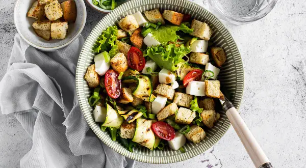 Deliziosa insalata di zucchine grigliate, pomodorini, cubetti di formaggio fresco e crostini croccanti, guarnita con erba cipollina.