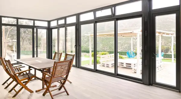 Comedor interior con mesa de madera y sillas, conectado a un jardín con porche acristalado. Precio a consultar.