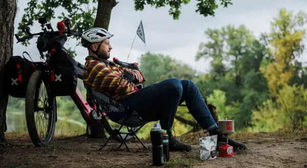 Mężczyzna odpoczywa podczas wycieczki rowerowej, gotując posiłek na kuchence turystycznej. Obok rower z sakwami.