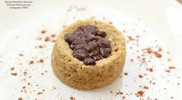Delicioso mug cake saludable con trozos de chocolate negro, espolvoreado con cacao en polvo sobre un plato blanco.