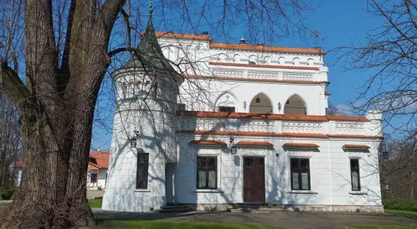 Biały pałac z wieżyczką i czerwonym dachem, otoczony drzewami. W oddali widać inne budynki. To piękny widok na **Radomyśl Wielki**.