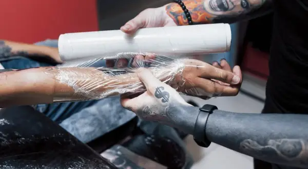 Man wrapping a fresh tattoo with plastic wrap. La curación es clave, y te preguntarás cuántos días hay que ponerse crema en un tatuaje para que quede perfecto.