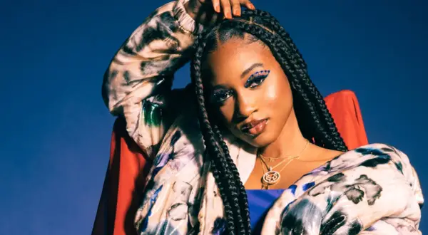 Nija Charles, with striking blue eye makeup and braids, poses confidently in a colorful jacket against a blue backdrop.