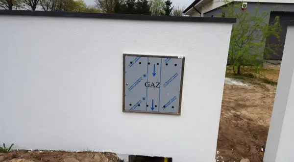 Metalowa skrzynka gazowa z napisem "GAZ" zamontowana na białym, otynkowanym murze.