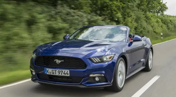 Błękitny mustang cabrio pędzi po krętej drodze wśród zieleni.