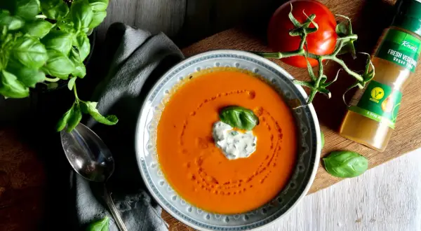 En skål med krämig tomatsoppa, gjord på krossade tomater, toppad med en klick gräddfil och basilika. En färsk tomat och en flaska Knorr Fresh Vegetable Vegan Fond ligger bredvid.