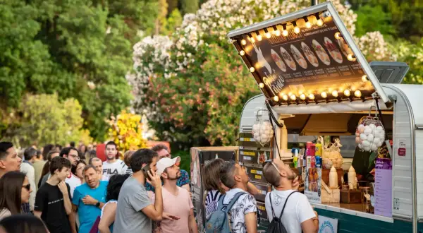 Gente disfrutando de food trucks en Zaragoza, un evento donde la gastronomía florece entre árboles y flores.