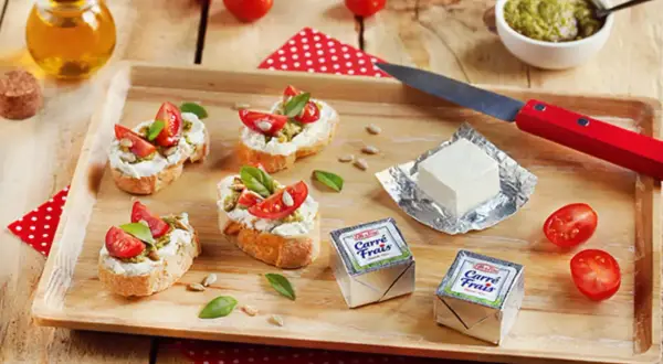 Bruschettas garnies de fromage frais, tomates et pesto, avec des emballages de Carré Frais le cendre et des tomates cerises sur un plateau en bois.