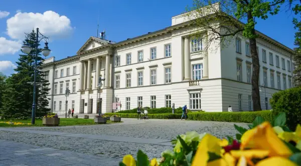 Biały budynek z kolumnami i zielenią, w tle błękitne niebo z chmurami.