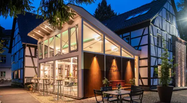 Abendstimmung an der buchmühle bergisch gladbach. Moderne Glasfront trifft auf Fachwerkhaus. Tische und Stühle laden zum Verweilen ein.