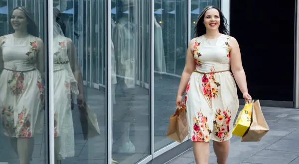 Uśmiechnięta kobieta w kwiecistej sukience plus size, z torbami zakupowymi w rękach, idzie obok witryny sklepowej.