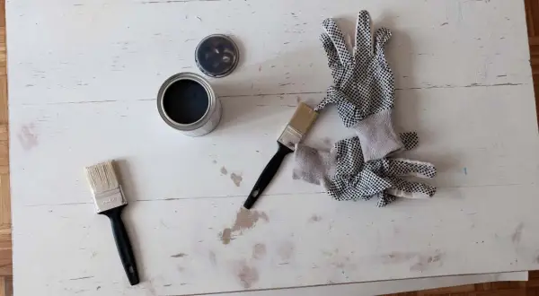 Narzędzia do renowacji stolika kawowego DIY: puszka czarnej farby, dwa pędzle i rękawice ochronne.