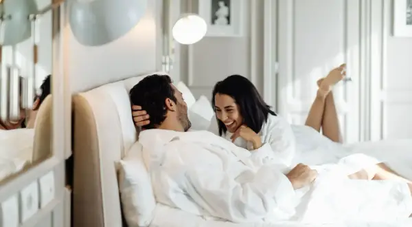 Ett lyckligt par i morgonrockar skrattar i en lyxig säng, kanske på ett hotell i St. Gallen.