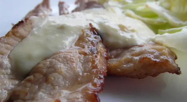 Tiernas lonchas de blanqueta de cerdo cubiertas con una cremosa salsa blanca, acompañadas de hojas verdes frescas.