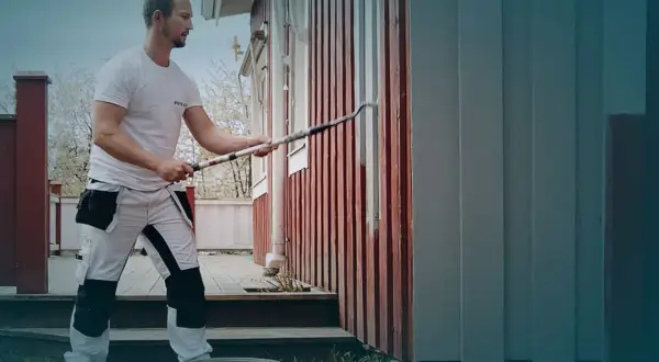 Man målar om huset med en lång roller. Det röda trähuset får nya gråa paneler.