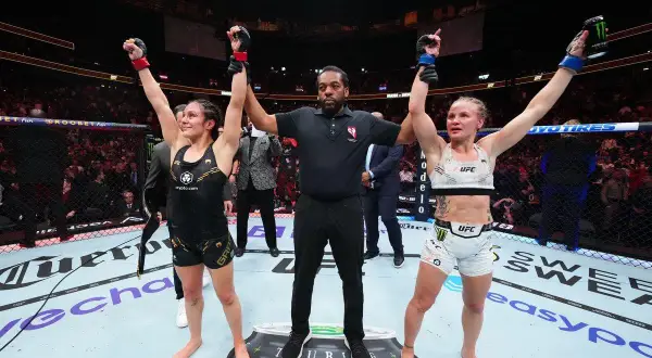 Two female fighters stand with arms raised, a referee between them. The outcome of their boxing draw is announced to the roaring crowd.