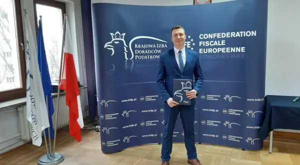 Mężczyzna w garniturze stoi przed niebieskim banerem z logo Krajowej Izby Doradców Podatkowych.