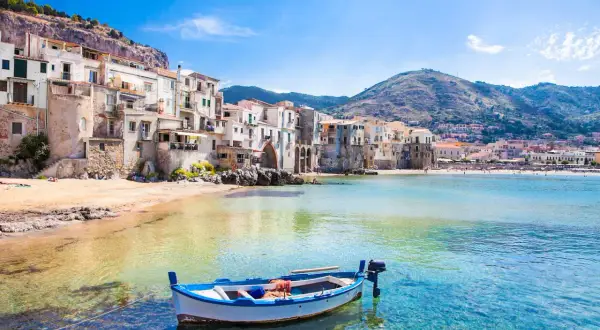 La **spiaggia più bella** di Cefalù, con case colorate a picco sul mare, una barca bianca e blu e acque cristalline.
