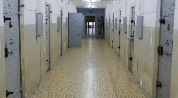 Korytarz więzienny z rzędami cel. Drzwi cel, niektóre z numerami jak 106 i 222, są zamknięte. W głębi widać kraty i otwarte drzwi.