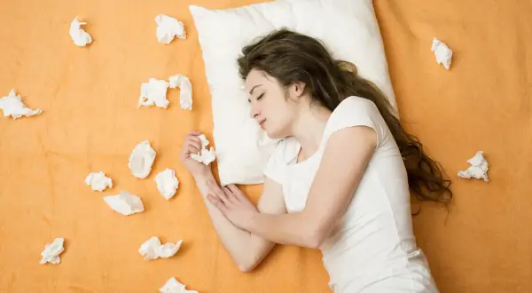 A young woman sleeps on a pillow surrounded by crumpled tissues, suggesting she might sneeze in her sleep.