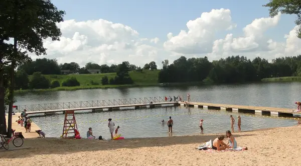 Letnie popołudnie na piaszczystej plaży nad jeziorem. Ludzie odpoczywają, kąpią się, a dzieci bawią się w wodzie. To typowy widok dla mazurskich miejscowości.