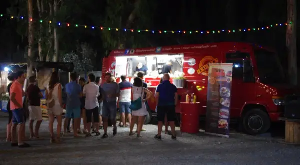 Gente disfrutando en un food trucks festival bajo luces de colores. Un camión rojo sirve comida mientras clientes esperan.