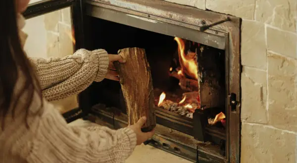 Une personne en pull ajoute une bûCHE dans un poêle à bois qui fume, porte ouverte. La chaleur des flammes réchauffe la pièce.
