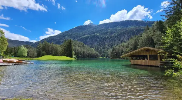 Ein idyllischer See mit türkisfarbenem Wasser, umgeben von grünen Hügeln und Wäldern. Ein Holzsteg mit Booten und ein kleines Holzhaus am Ufer laden zum Verweilen ein. Perfekt für einen blindsee trail.