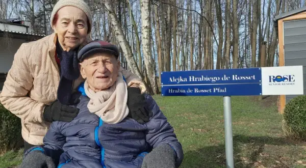 Uśmiechnięta para seniorów pozuje przy tablicy "Alejka Hrabiego de Rosset" w dom seniora Rotmanka.