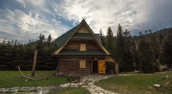 Drewniana bacówka w otoczeniu lasu, z kamienną ścieżką i chmurami na niebie.