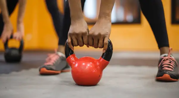 Dłonie zaciskają uchwyt czerwonej kettlebell, gotowe do treningu crossfit. W tle widać inne osoby ćwiczące.