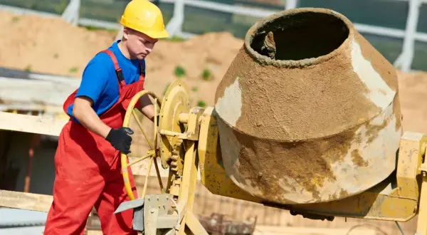 Robotnik w żółtym kasku i czerwonym ogrodniczkach obsługuje betoniarkę. Kluczowe są tu beton bez żwiru proporcje.