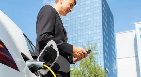 Uomo in giacca e cravatta controlla il telefono mentre la sua auto elettrica si ricarica, pianificare viaggio auto elettrica è facile con la tecnologia.