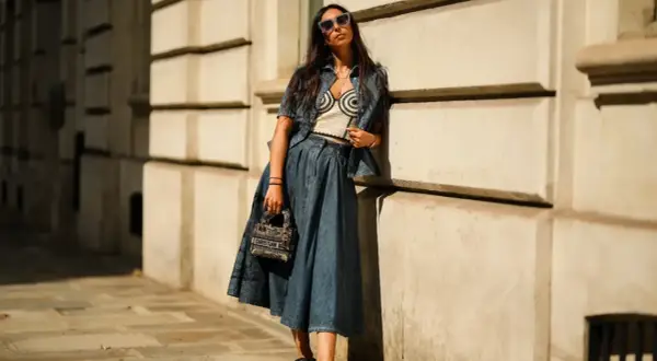 Mujer con gafas de sol posa apoyada en una pared, luciendo un conjunto vaquero y una falda vaquera larga.