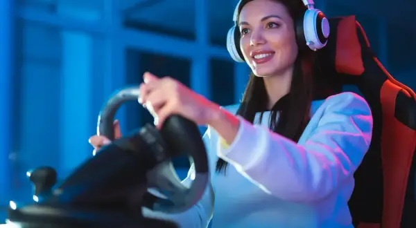 Uśmiechnięta kobieta w słuchawkach gra w wyścigi na kierownicy. Motoryzacja i rozrywka łączą fanów szybkich aut i gier online.