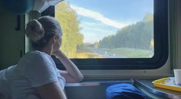 Frau blickt aus dem Fenster eines Nachtzuges nach Italien. Die Landschaft zieht vorbei, Bäume und eine Straße sind zu sehen.