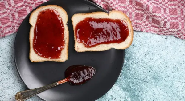Zwei Scheiben Toast mit leckerer Jostabeeremarmelade und ein Löffel voll davon auf einem schwarzen Teller.