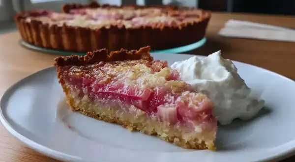 En bit härlig rabarberpaj glutenfri med en klick vispad grädde. Hela pajen syns i bakgrunden.