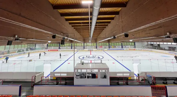 Eishalle Wiehl mit Eisfläche, Banden und Tribüne. Einige Eisläufer sind auf dem Eis zu sehen.